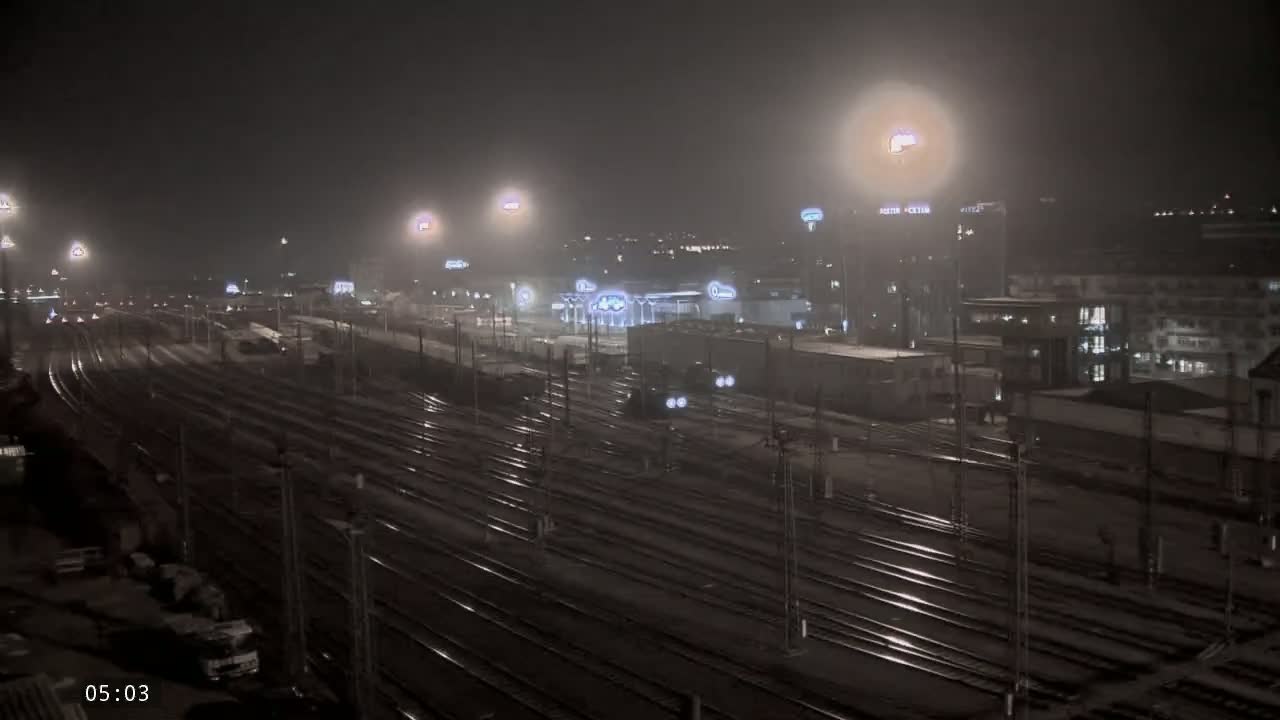 A nighttime view of a train station with many railway tracks and buildings under foggy conditions.