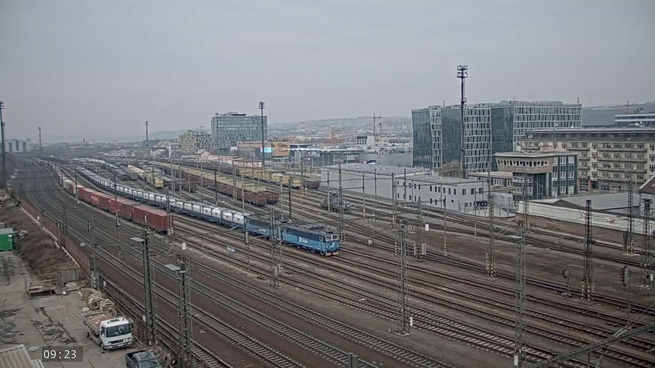 A long freight train is moving through a large railway yard on an overcast day, with modern buildings in the background.
