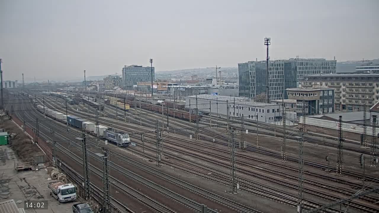 A sprawling railway yard with numerous train cars and tracks is visible under an overcast sky, with modern buildings in the background.