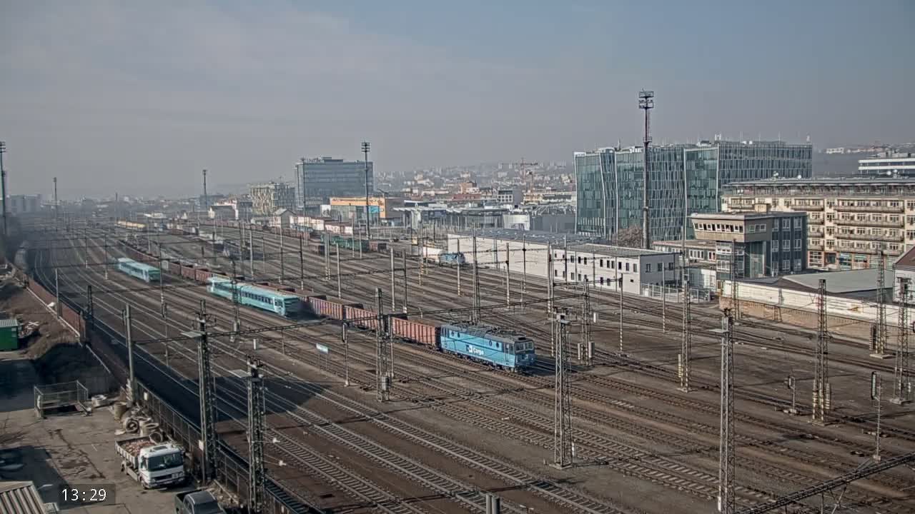 A sprawling railway yard with multiple trains and freight cars, under a hazy, sunny sky, is situated next to a city with modern and older buildings.