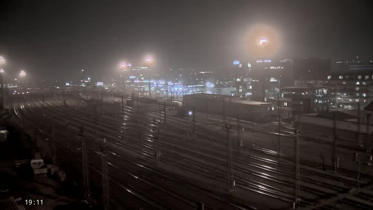 A nighttime view of a foggy train yard with numerous tracks and buildings in the background.