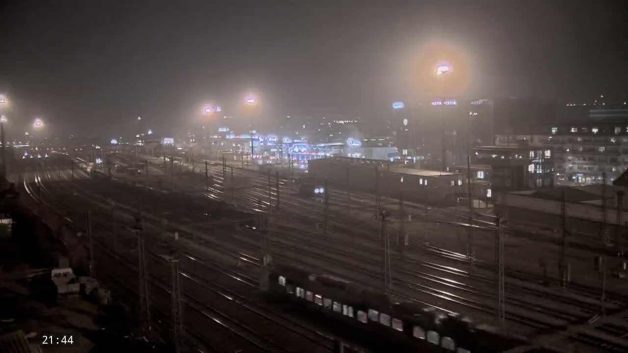 A long nighttime view of a train station with many train tracks, buildings, and lights in foggy conditions.