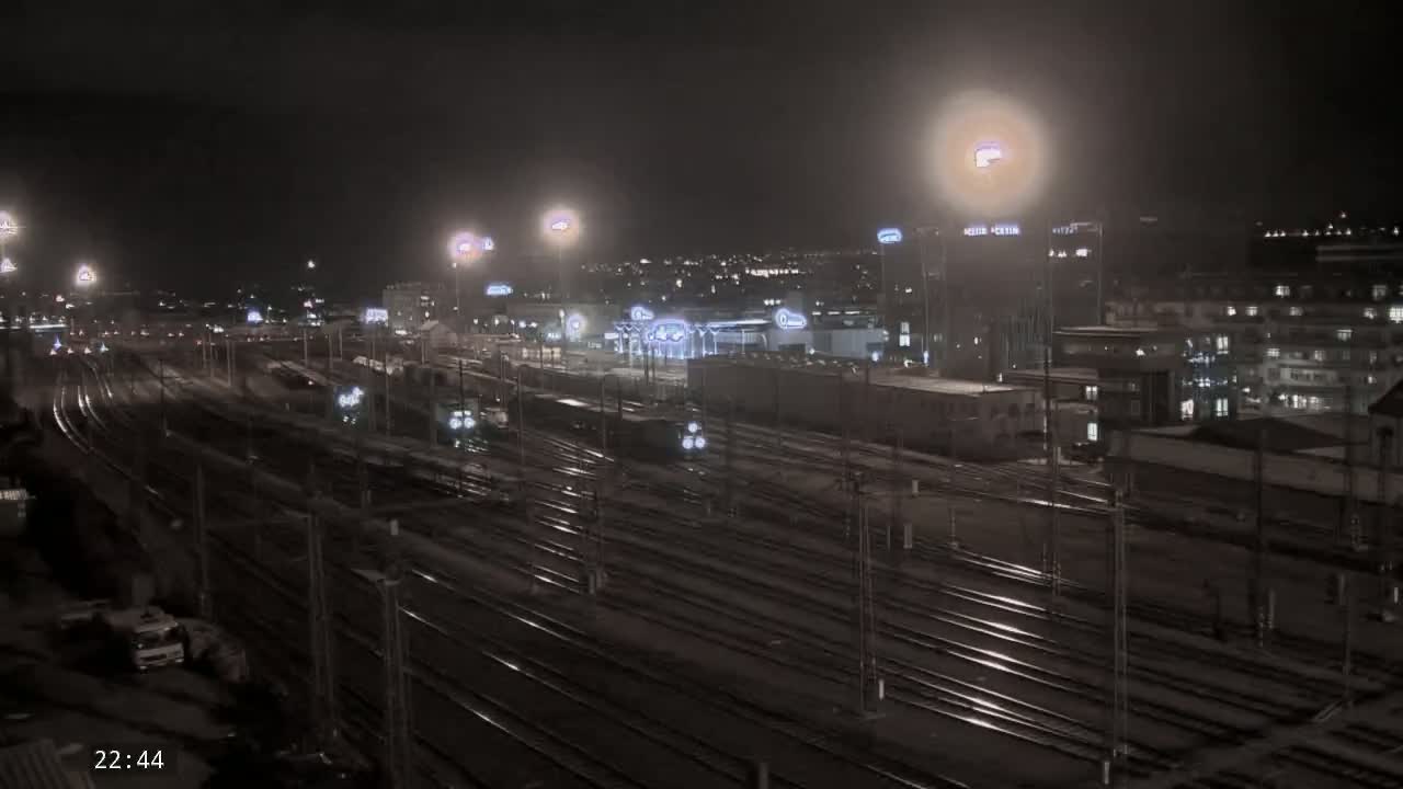 A nighttime view of a railway yard with numerous train tracks, some trains, and buildings in the background under artificial light.