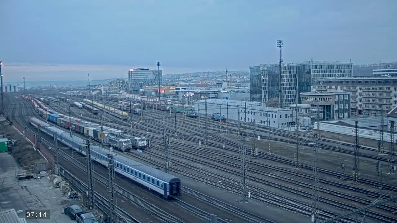 A long freight train and passenger train travel along numerous railway tracks in a city under an overcast sky.