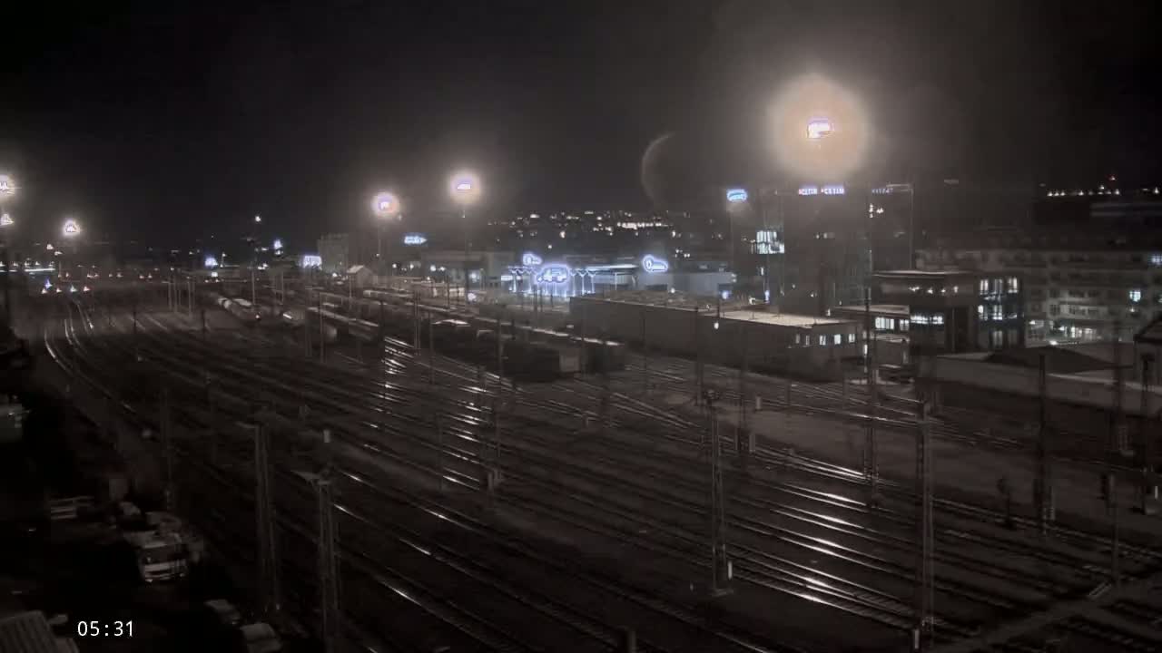 A nighttime view of a large railway yard with many train tracks, illuminated by artificial lights, and buildings in the background under a dark, clear sky.