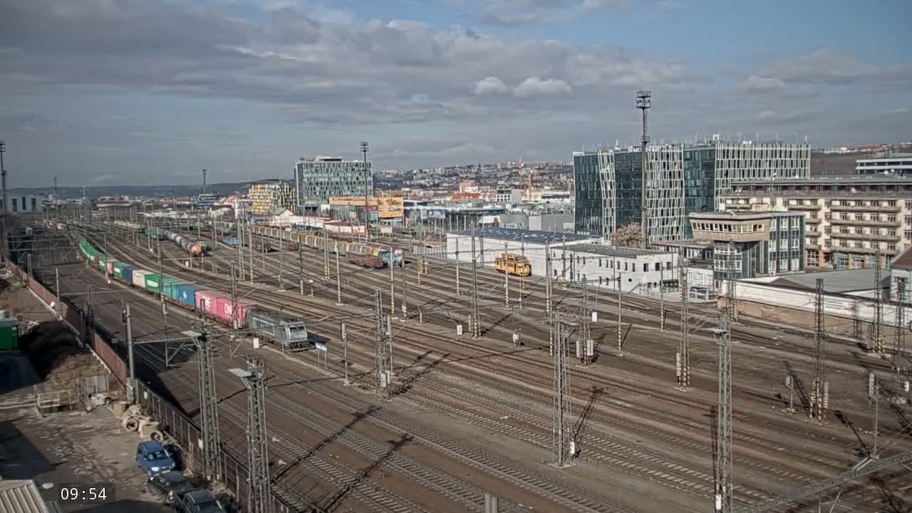 A sprawling railway yard with numerous tracks, trains carrying cargo containers, and surrounding buildings under a partly cloudy sky.