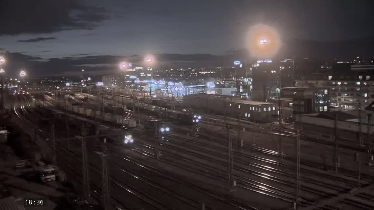 A nighttime view of a sprawling rail yard with numerous trains and tracks, surrounded by city buildings under a partly cloudy sky.
