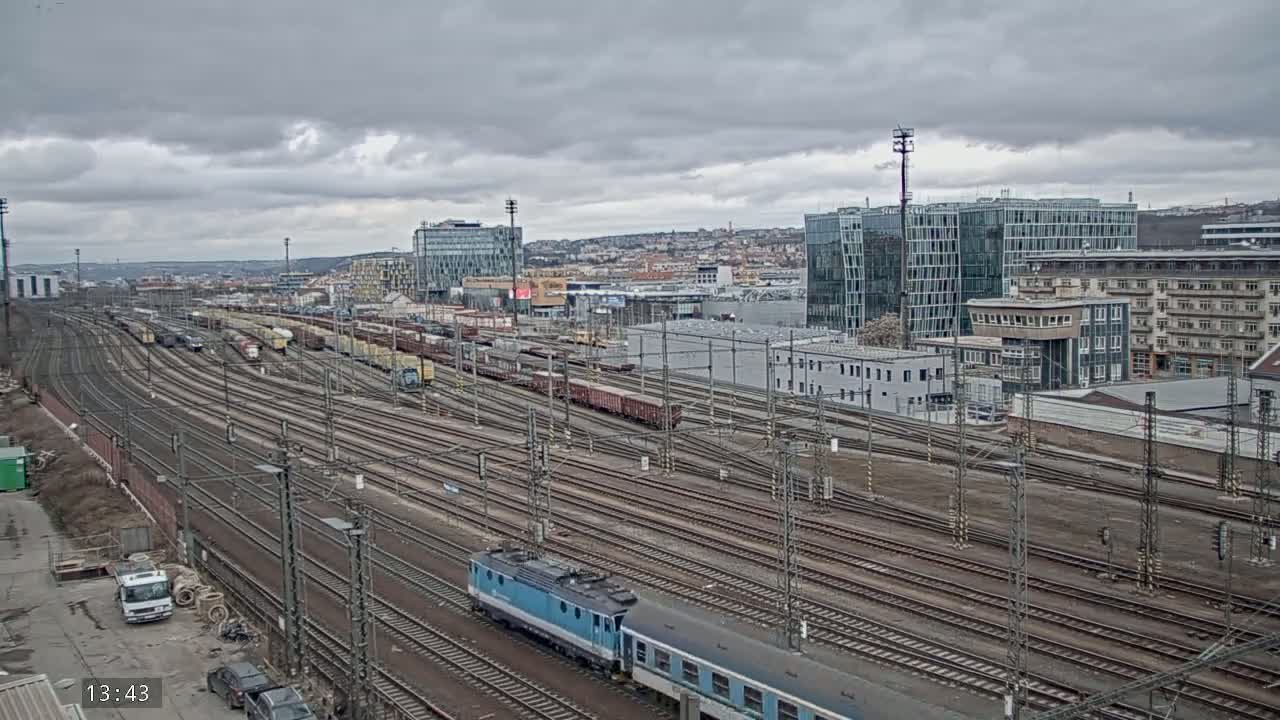 A sprawling railway yard with numerous tracks and freight cars, under a cloudy sky, is adjacent to a city with modern and older buildings.