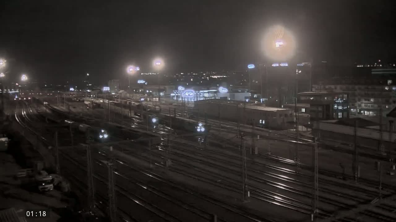 A nighttime view of a train yard with many tracks and several buildings in the background, under hazy conditions.
