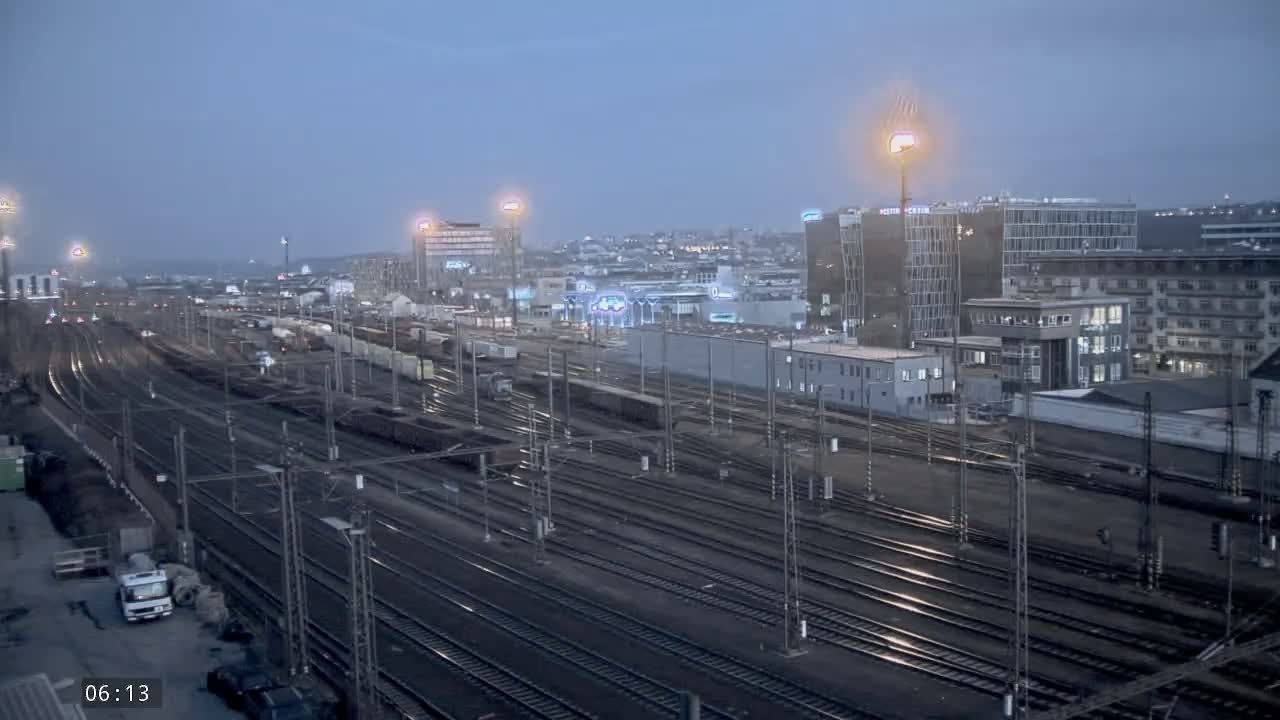 A sprawling, dimly lit railway yard with numerous train tracks and freight cars is situated beneath a twilight sky next to several multistory buildings.