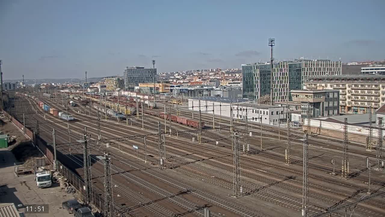 A large railway yard with numerous train cars and tracks stretches out under a clear blue sky, beyond which are modern and older buildings.