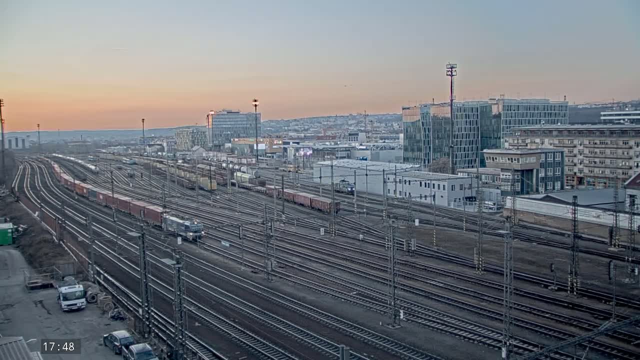 A large railway yard with numerous train cars and tracks is visible under a dusky pink sunset sky.