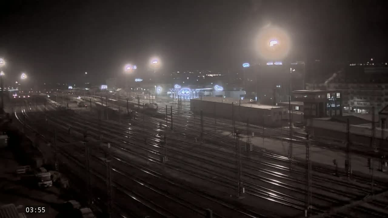 A nighttime view of a snow-covered train yard with numerous tracks extending into the distance, under hazy, dimly lit conditions.