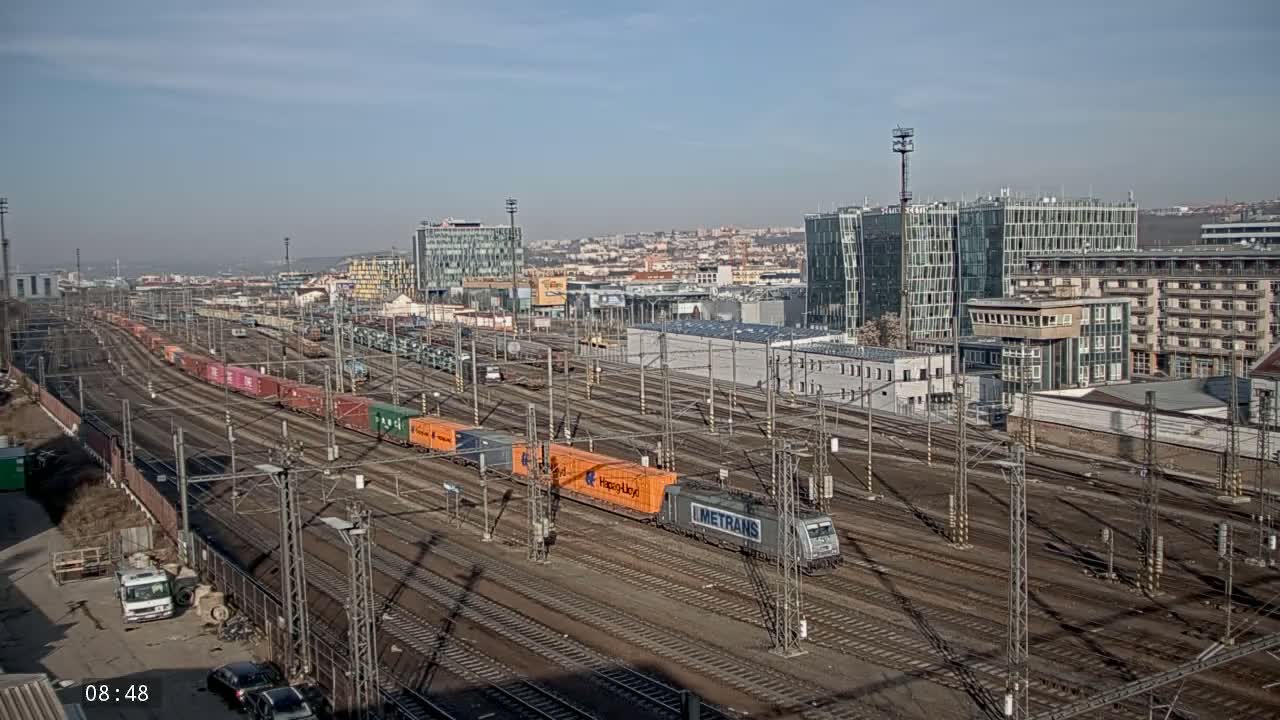 A freight train is moving through a large railway yard on a sunny day, with numerous other train cars and tracks visible, and city buildings in the background.