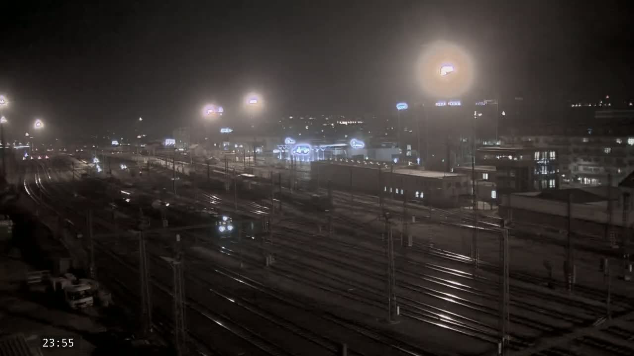 A nighttime view of a train station with many tracks and surrounding buildings under hazy, possibly snowy conditions.