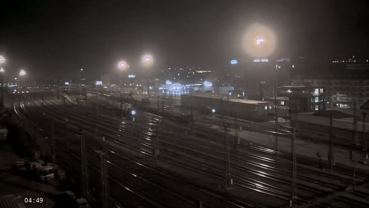 A nighttime, hazy view of a train yard with numerous tracks and surrounding buildings under artificial lights.