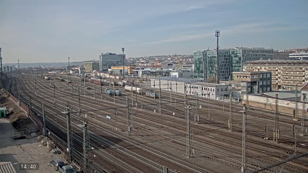 A sprawling railway yard with numerous tracks, freight cars, and buildings in the background under a mostly clear sky.