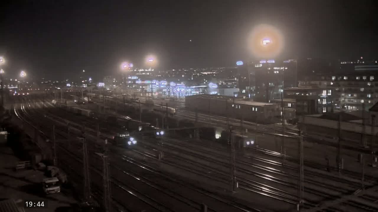 A nighttime view of a train yard with several trains and buildings illuminated under hazy conditions.