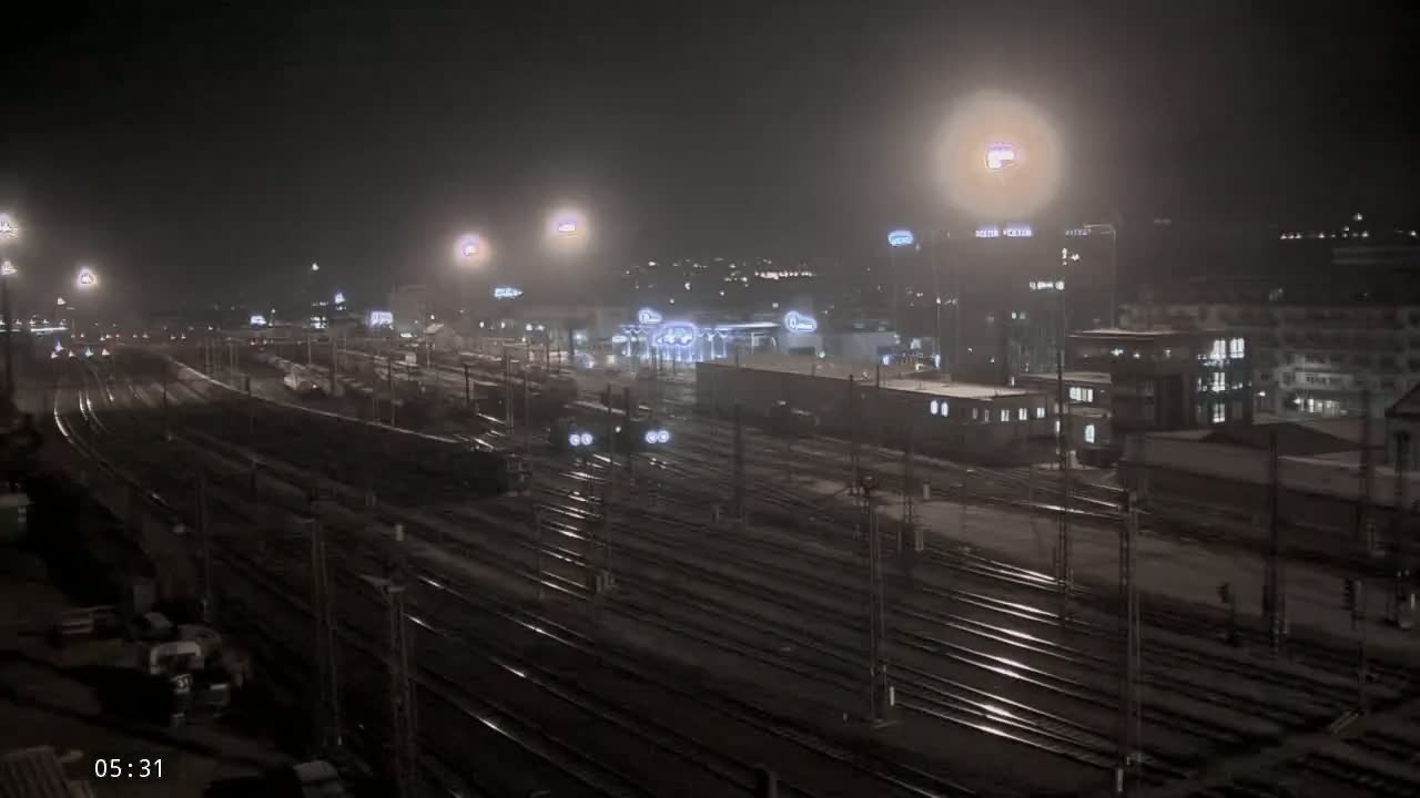 A nighttime view of a snowy train yard with numerous tracks and buildings under hazy, illuminated conditions.