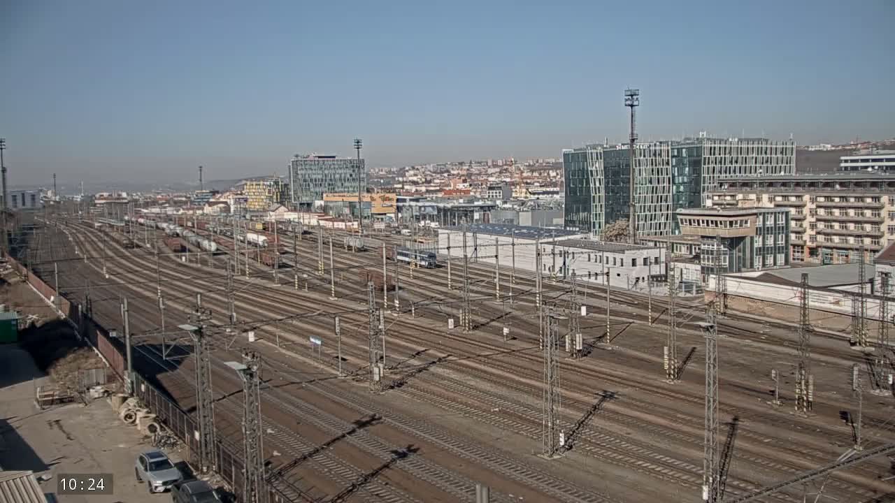 A large railway yard with numerous tracks and freight cars extends into the distance under a clear blue sky, bordered by modern and older buildings.