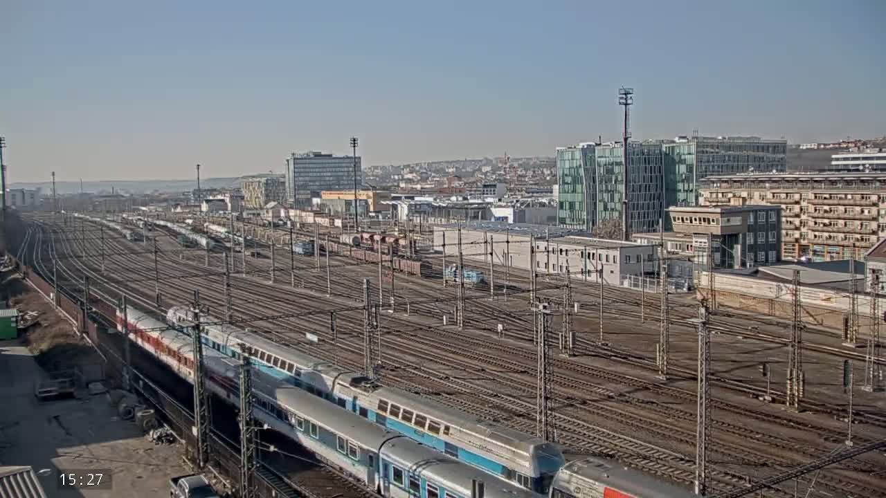 A sunny day reveals a large train yard with numerous tracks, several trains, and a cityscape in the background.