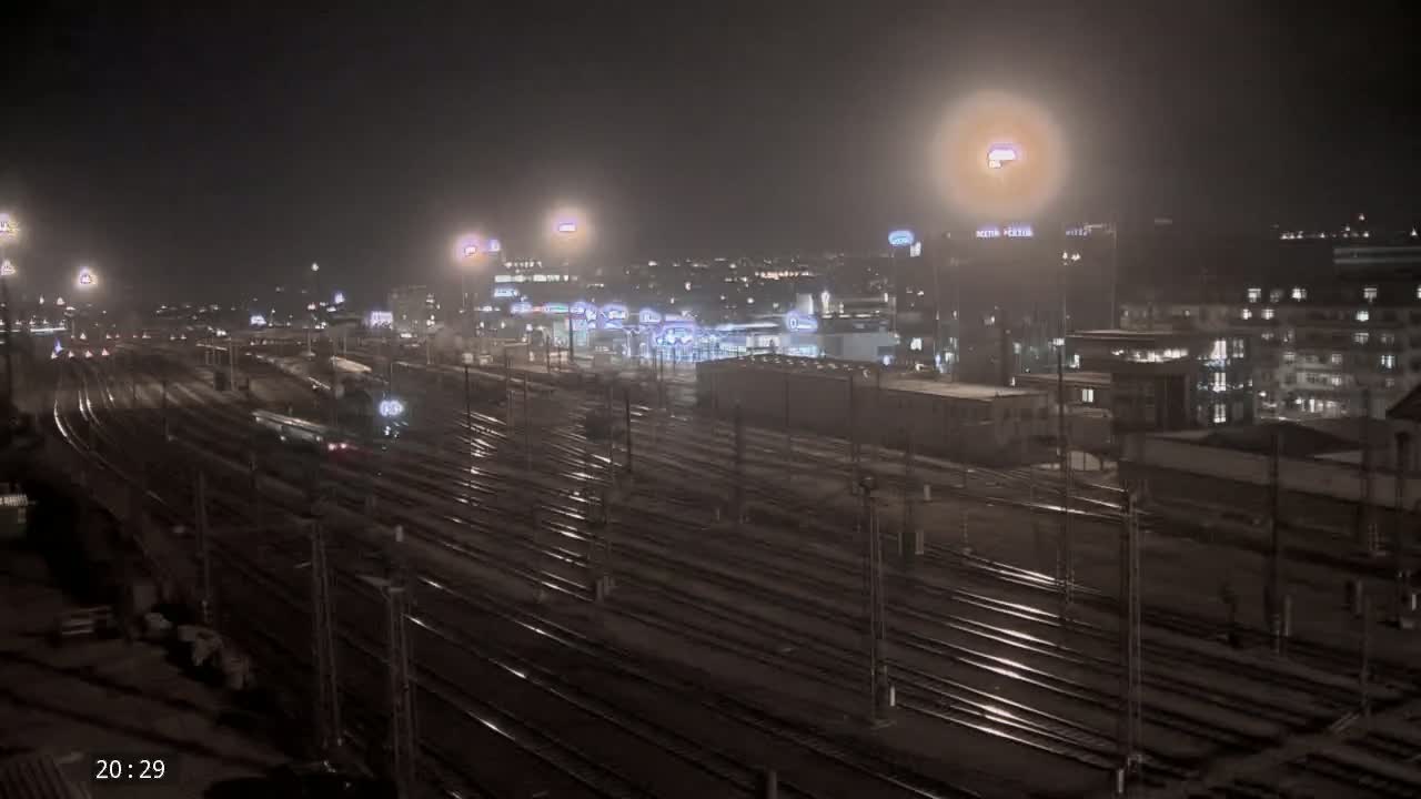 A nighttime view of a train station with numerous train tracks, illuminated by lights, and a hazy atmosphere.