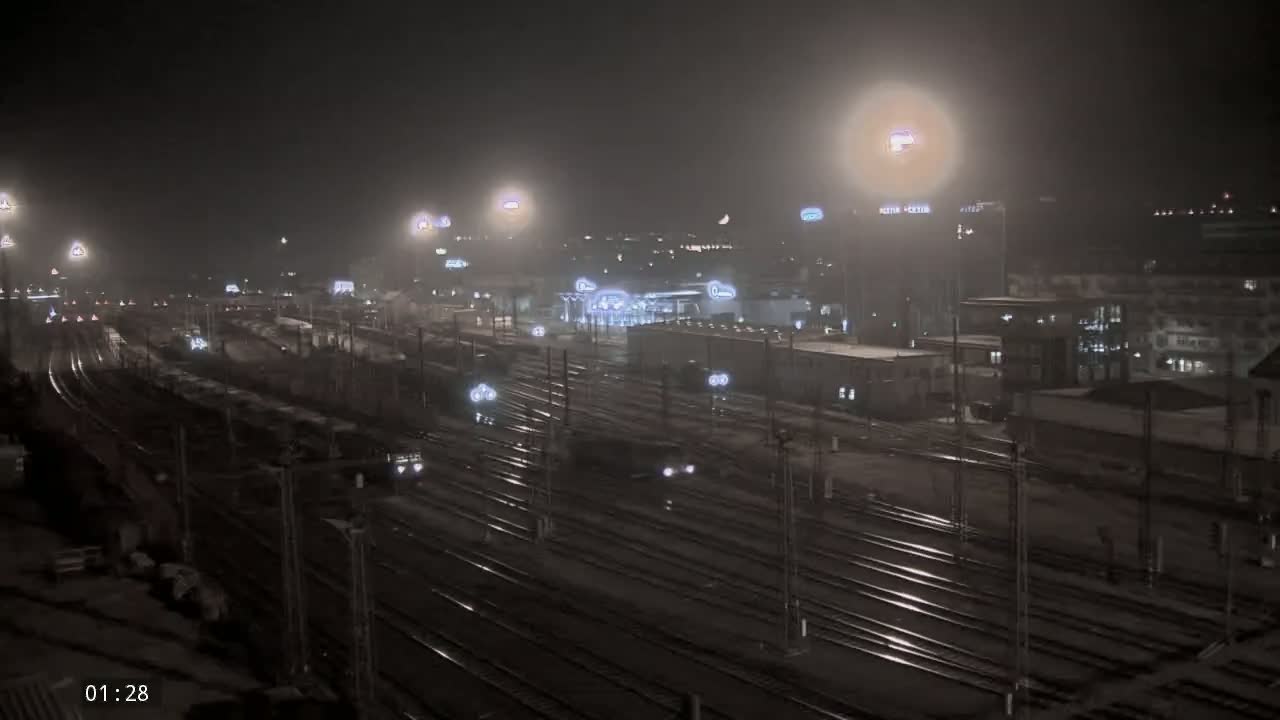 A nighttime view of a snow-covered train yard with multiple tracks and several buildings in the background under hazy, dimly lit conditions.