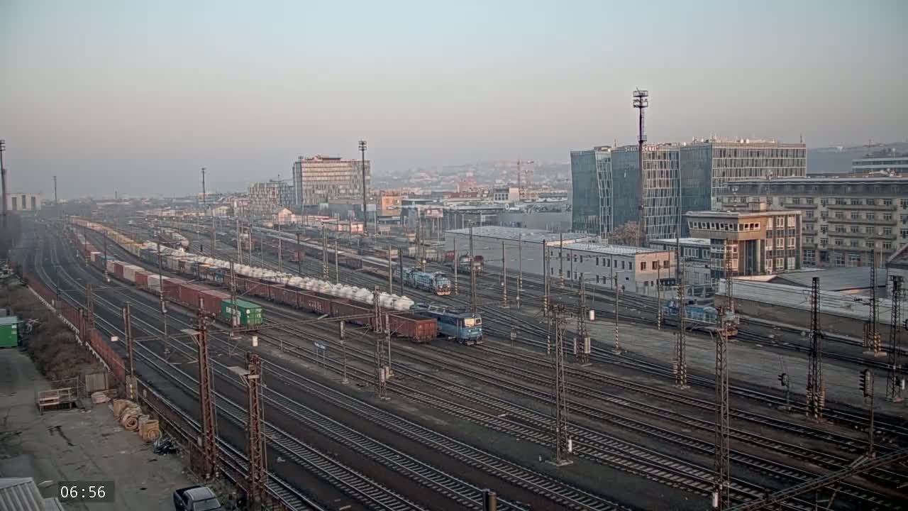 A long freight train is traveling through a large railway yard on a hazy morning, with several buildings in the background.