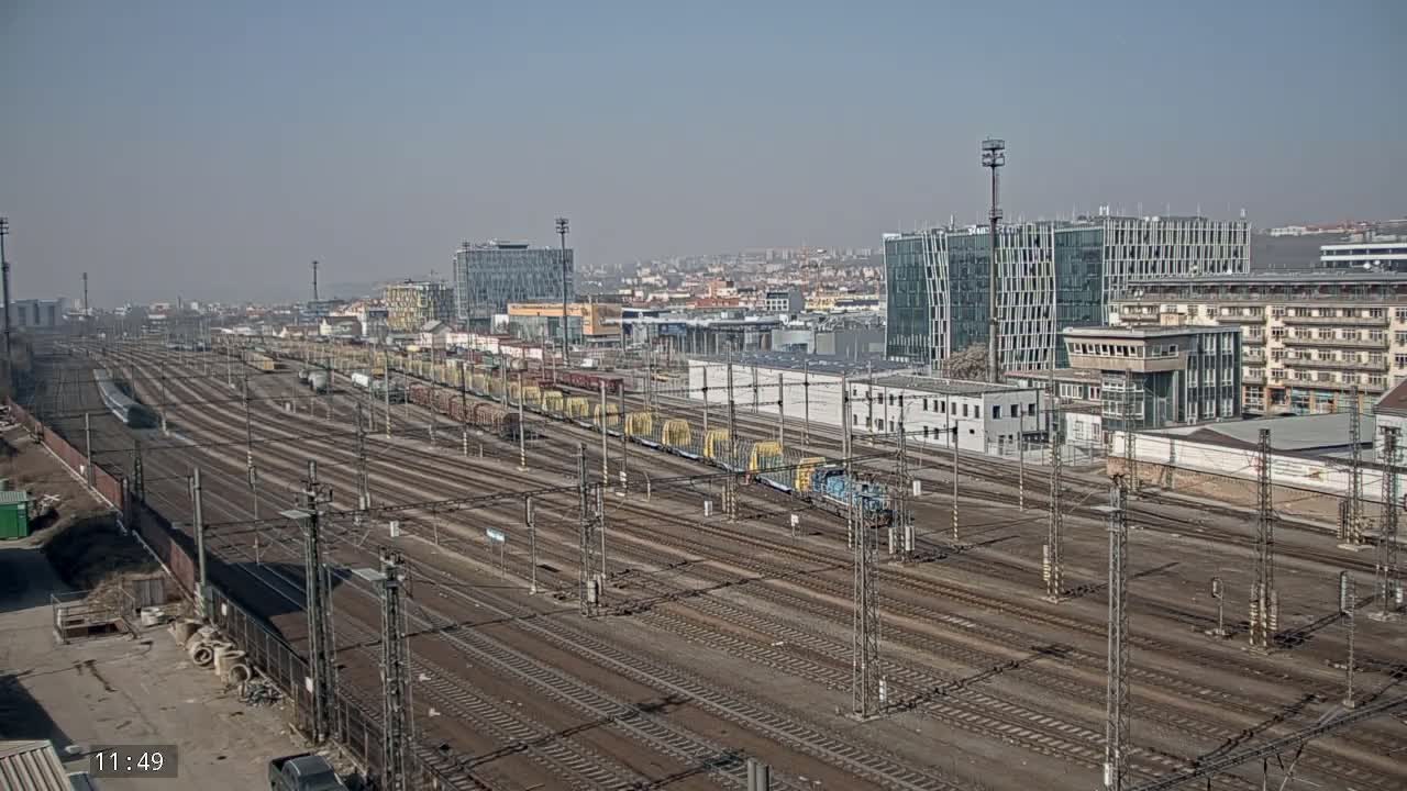 A large railway yard with many parallel tracks and several freight trains, under a clear, sunny sky, is situated near a city with modern and older buildings.