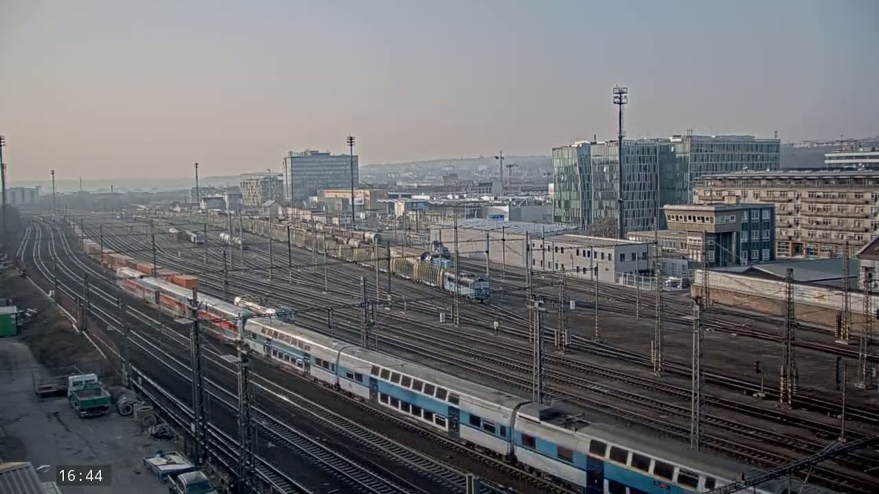 A long passenger train travels through a sprawling rail yard on a hazy day, past numerous other trains and industrial buildings.