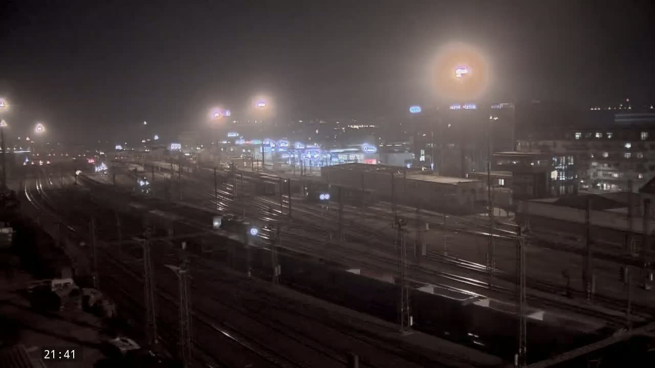 A nighttime view of a foggy train yard with numerous tracks and some buildings in the background.