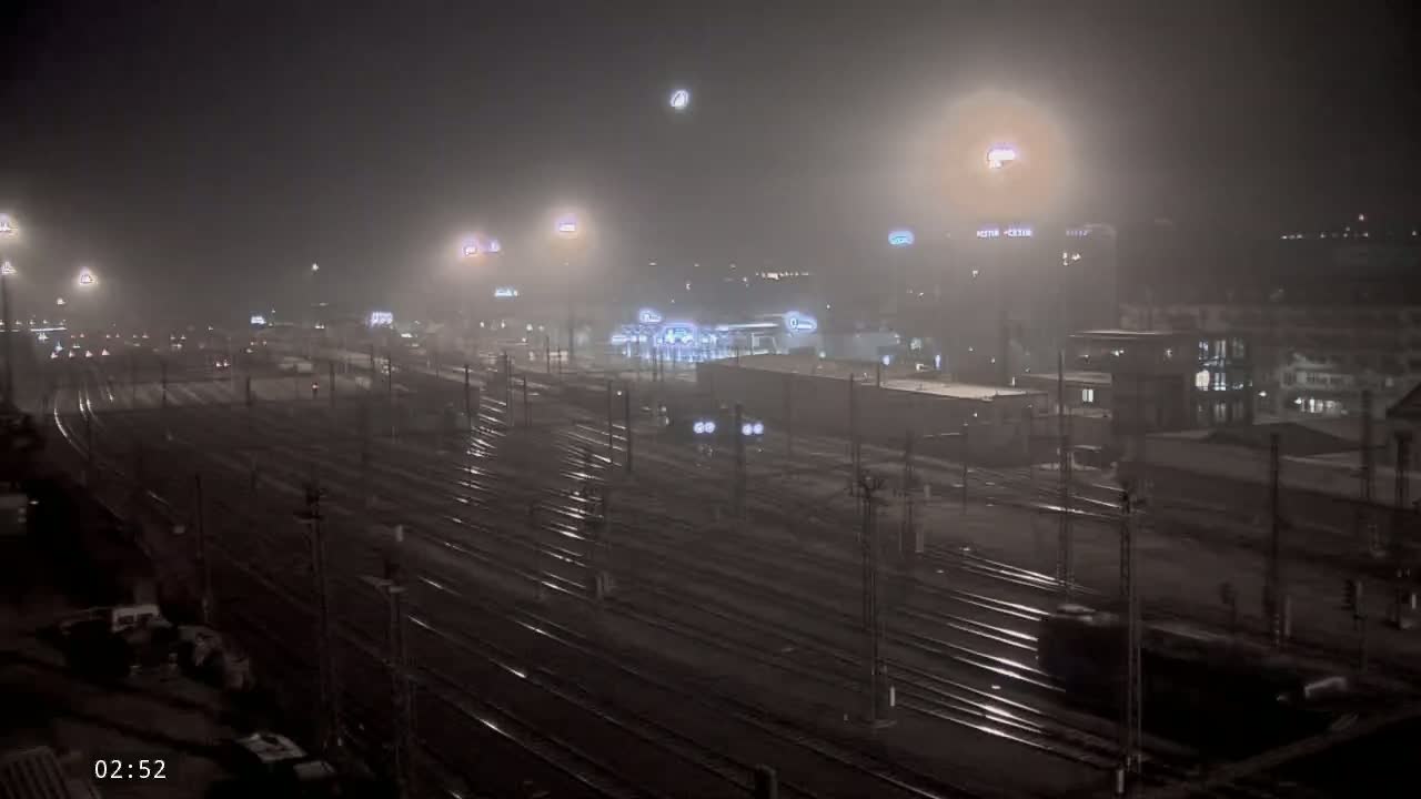 A foggy night scene of a train yard with numerous tracks and dimly lit buildings in the background.