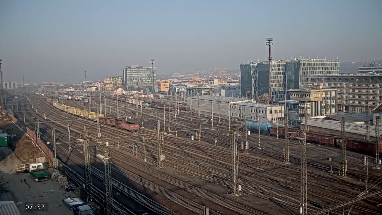 A hazy, sunny day reveals a sprawling railway yard filled with numerous train cars and tracks, adjacent to a cityscape of modern and older buildings.