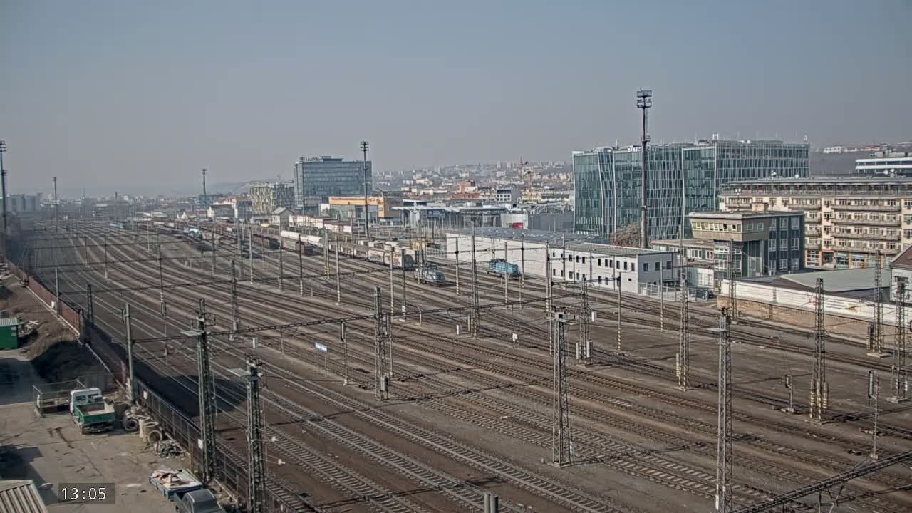 A sprawling railway yard with numerous tracks extends into the distance under a clear, sunny sky, bordered by modern and older buildings.