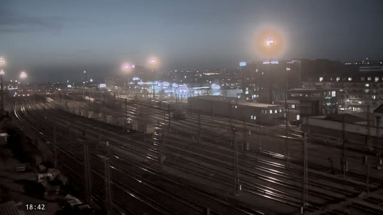 A nighttime view of a sprawling railway yard with numerous tracks, adjacent to a city skyline under hazy, dim lighting.