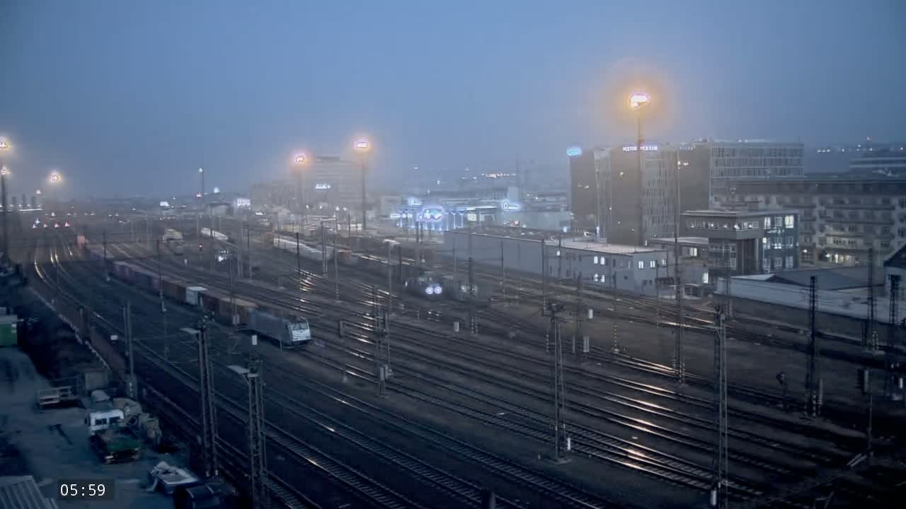 A foggy, early morning view of a large railway yard with multiple train cars and tracks, surrounded by city buildings.