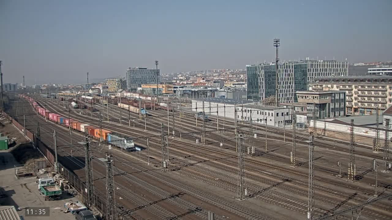 A sprawling railway yard with numerous train tracks, several trains, and a cityscape in the background under a clear, sunny sky.