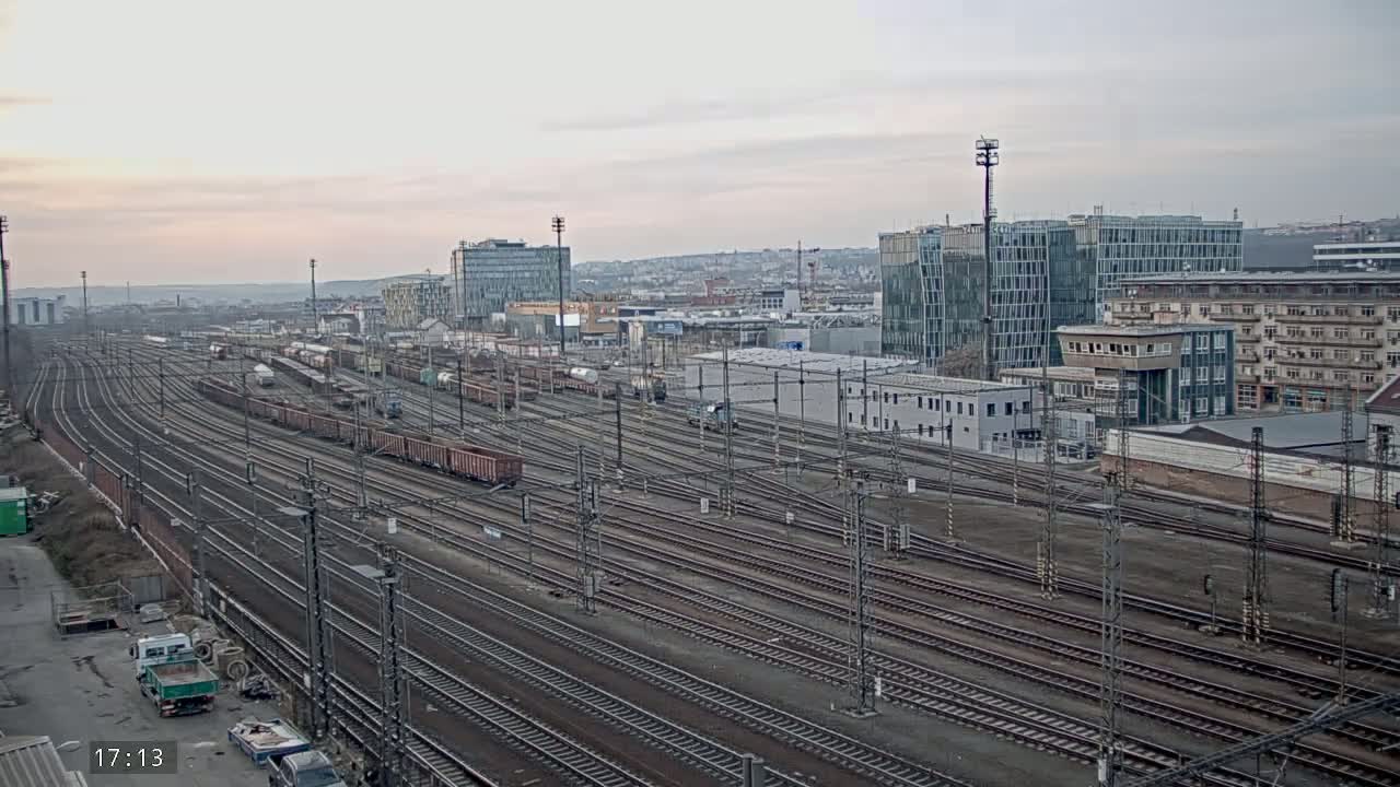 Numerous train tracks extend into the distance, past freight cars and city buildings under an overcast sky.