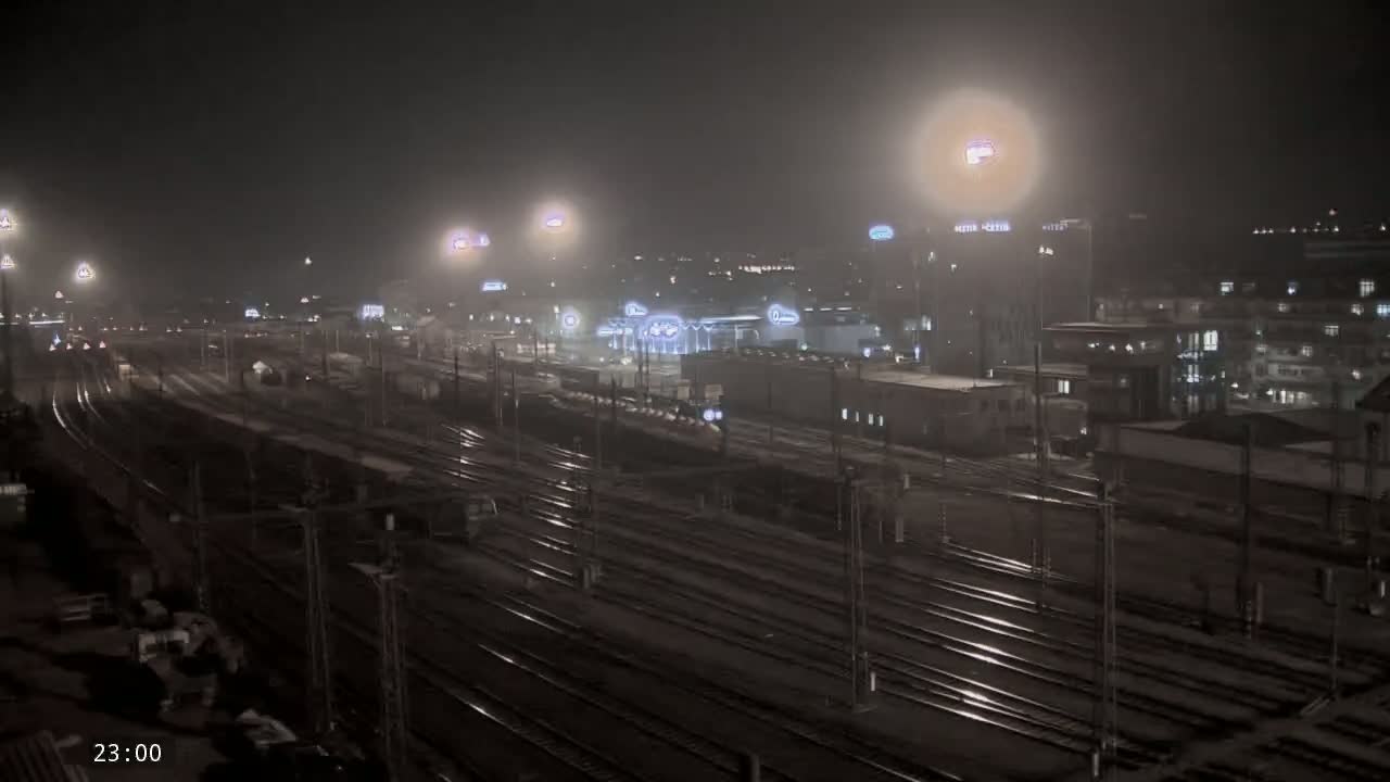 A nighttime view of a train yard with numerous tracks under hazy or foggy conditions, surrounded by buildings with illuminated windows.