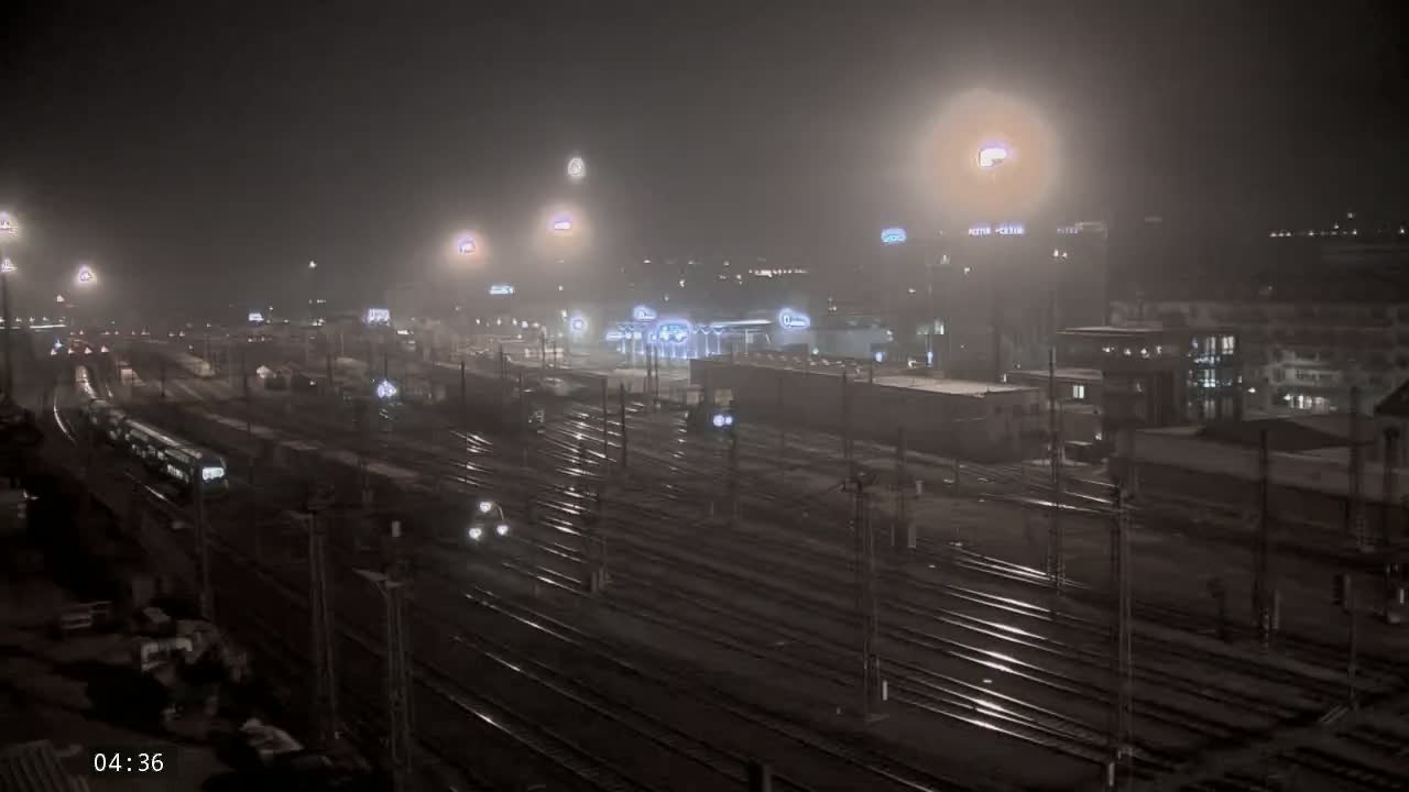 A foggy night scene shows a train station with multiple tracks, trains, and buildings under bright lights.