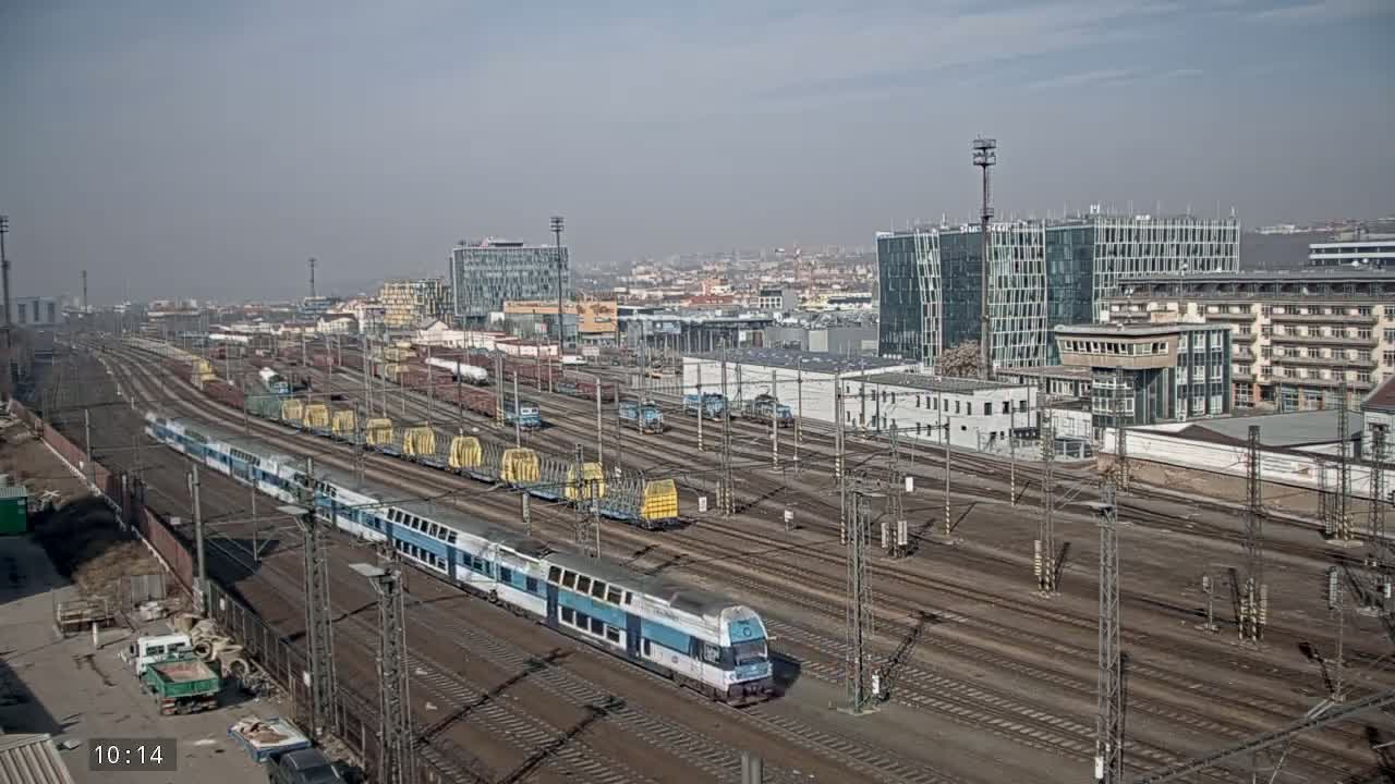 A long passenger train passes through a large, sprawling train yard on a hazy, sunny day, with numerous freight cars and modern buildings in the background.