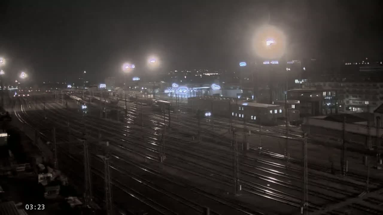 A nighttime view of a sprawling, wet train yard illuminated by numerous lights under a hazy sky.