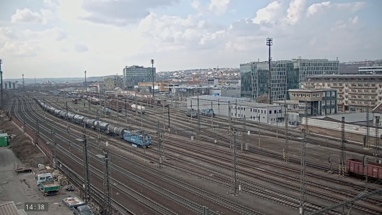 A sprawling railway yard with numerous train tracks and several trains, including one long freight train, under a partly cloudy sky.
