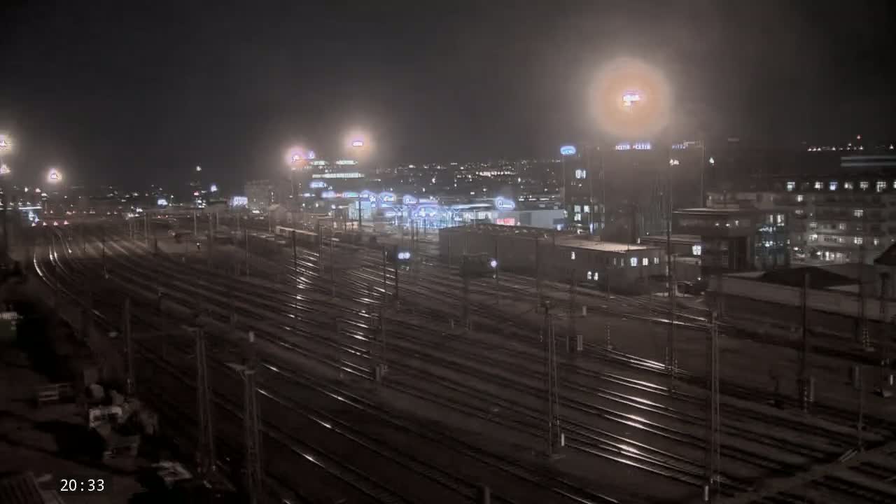 A nighttime view of a vast railway yard with many tracks, illuminated by artificial lights, beside a city skyline under a hazy sky.