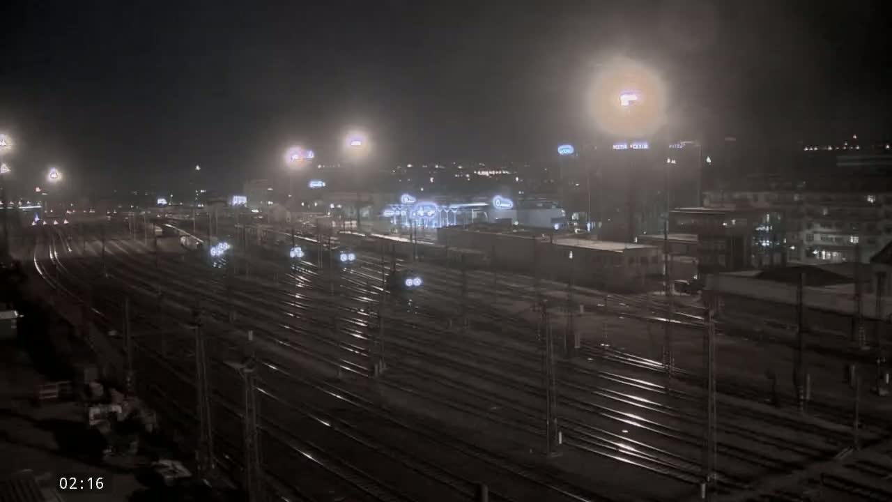 A nighttime, dimly lit view of a vast train yard with numerous tracks and trains, surrounded by buildings under hazy or foggy conditions.
