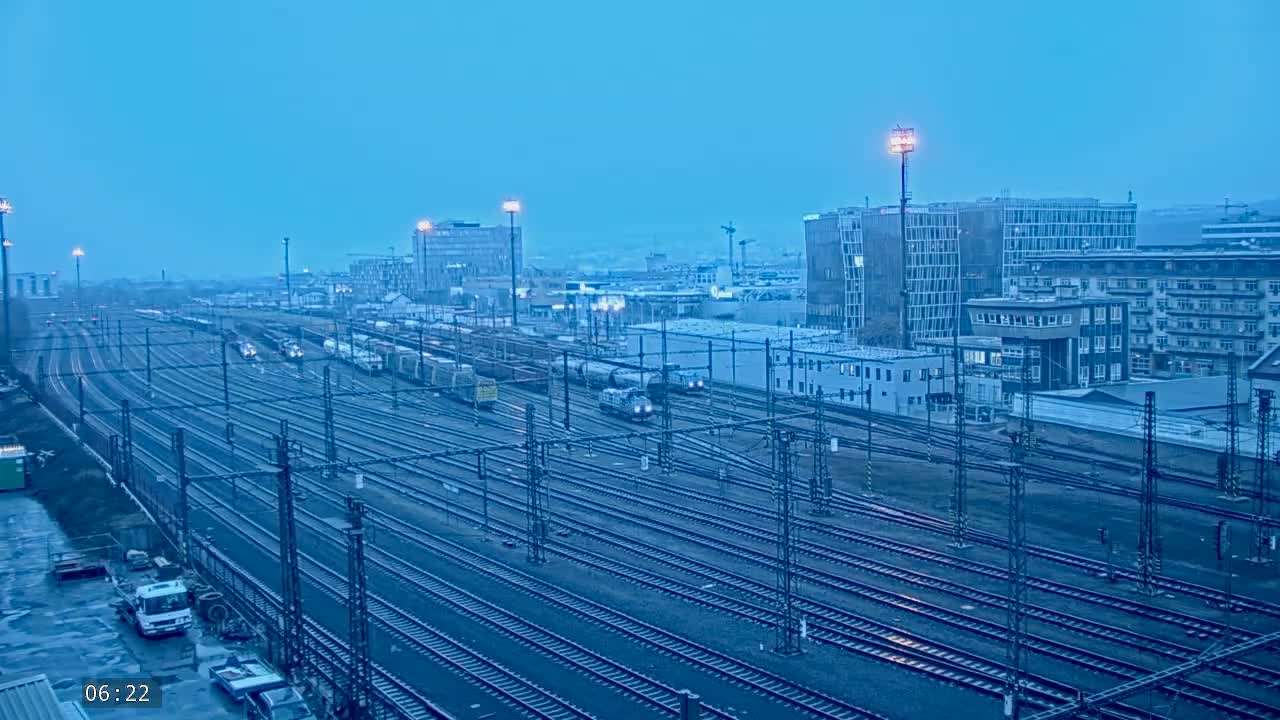 Under a twilight blue sky, numerous train cars and locomotives sit on a vast network of train tracks in a city with several buildings visible in the background.