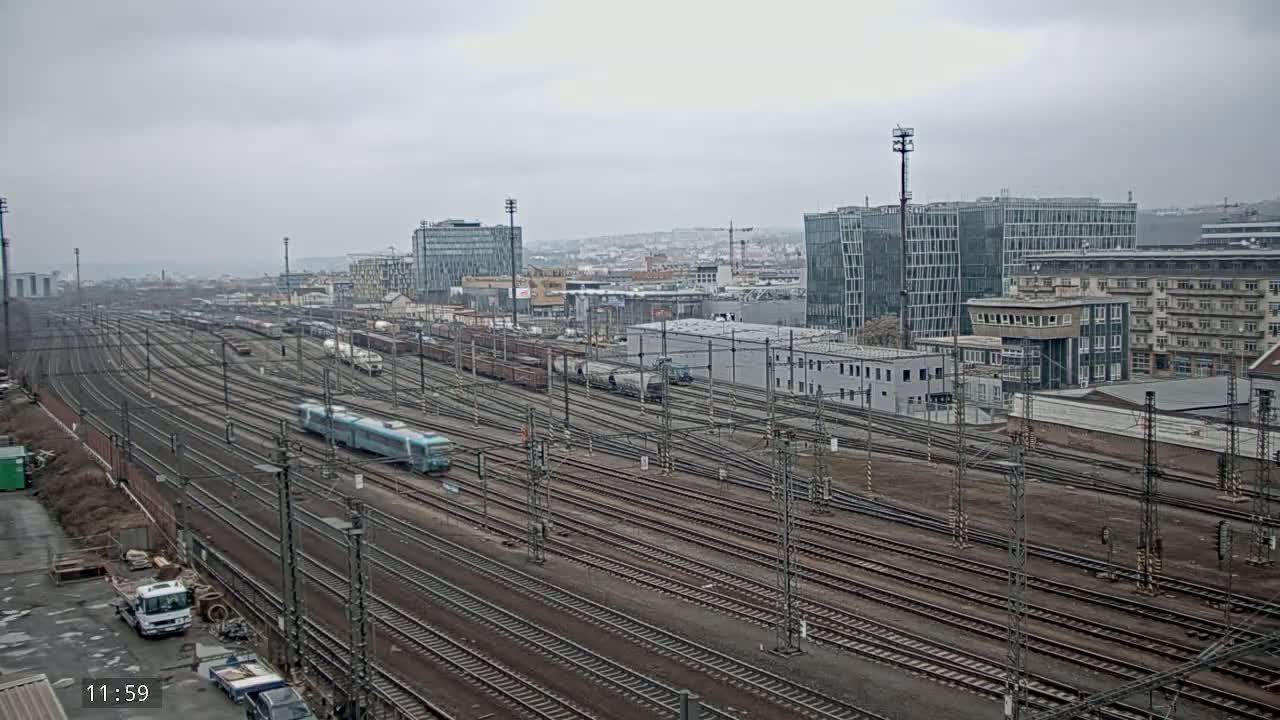 A wide view of numerous train tracks in a city with overcast skies, several trains visible on the tracks, and buildings in the background.