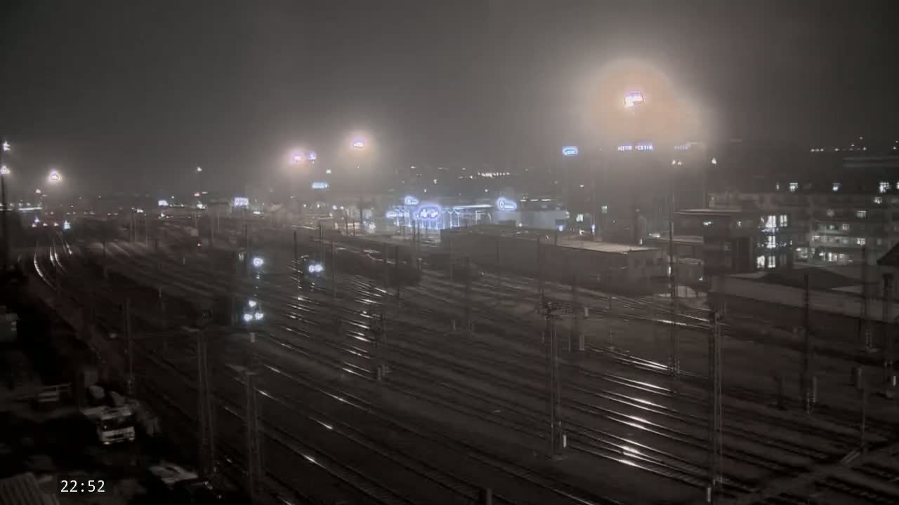 A foggy night scene shows a train yard with many railway tracks and several trains, illuminated by lights.
