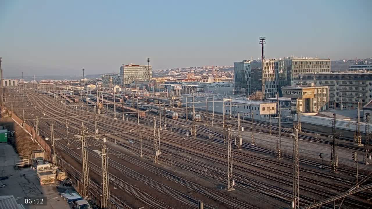 A sprawling railway yard with numerous train cars and tracks extends towards a city skyline under a clear, bright sky.