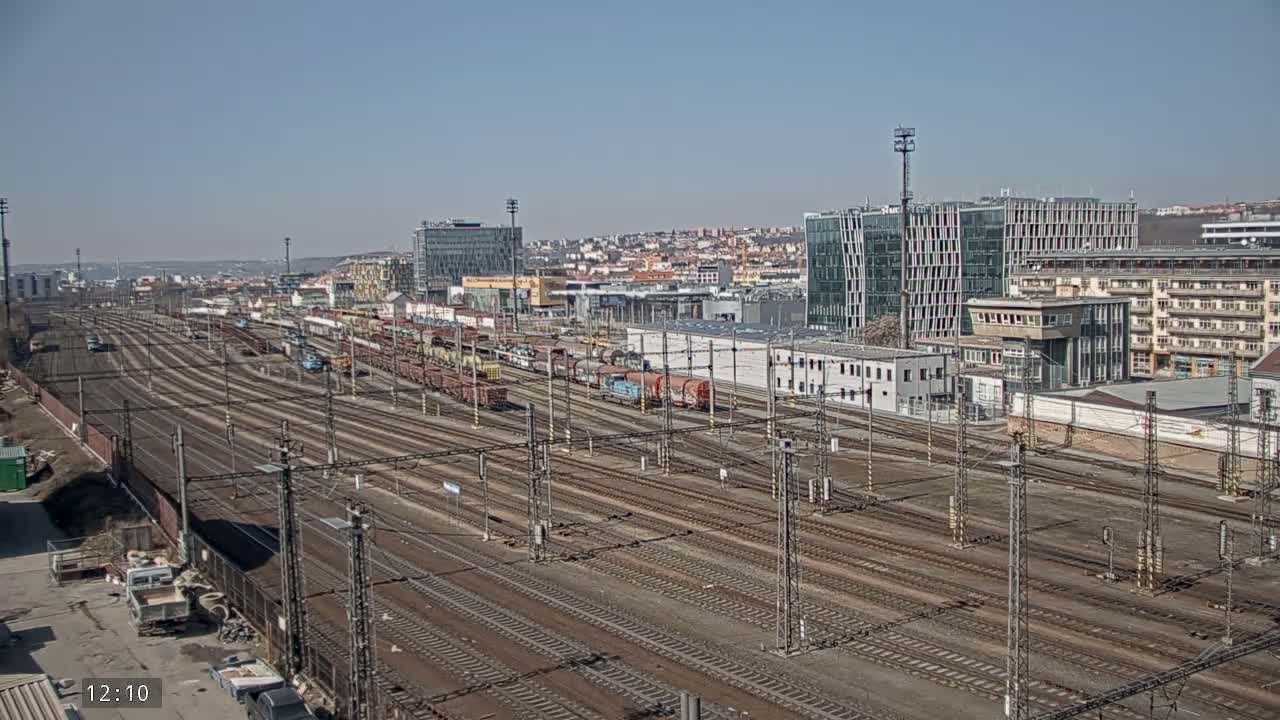 A wide view of a large railway yard with numerous train cars and tracks, under a clear sunny sky, is surrounded by buildings of varying heights and architectural styles.
